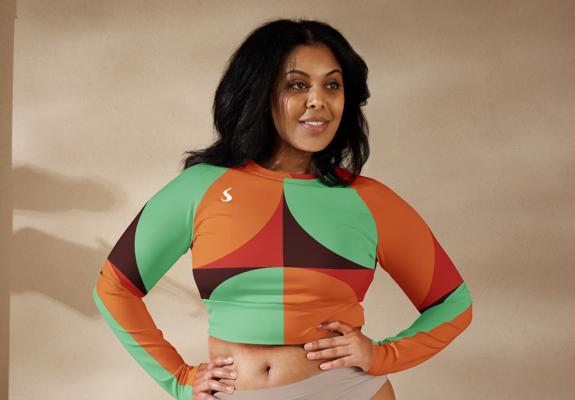 Crop top de sport Hot Focus à manches longues, motif géométrique au couleur vert d'eau, brun, rouge et orange, conçu pour allier confort et style lors des entraînements. Photo porté par le mannequin taille L debout, bras sur les hanches.