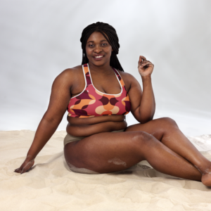 Brassière de sport Sweat Hold Up en nylon Econyl, douce et respirante, avec dos nageur pour un maintien léger. Disponible dans plusieurs motifs. Photo du produit porté par une mannequin plus size, elle est assise sur du sable.